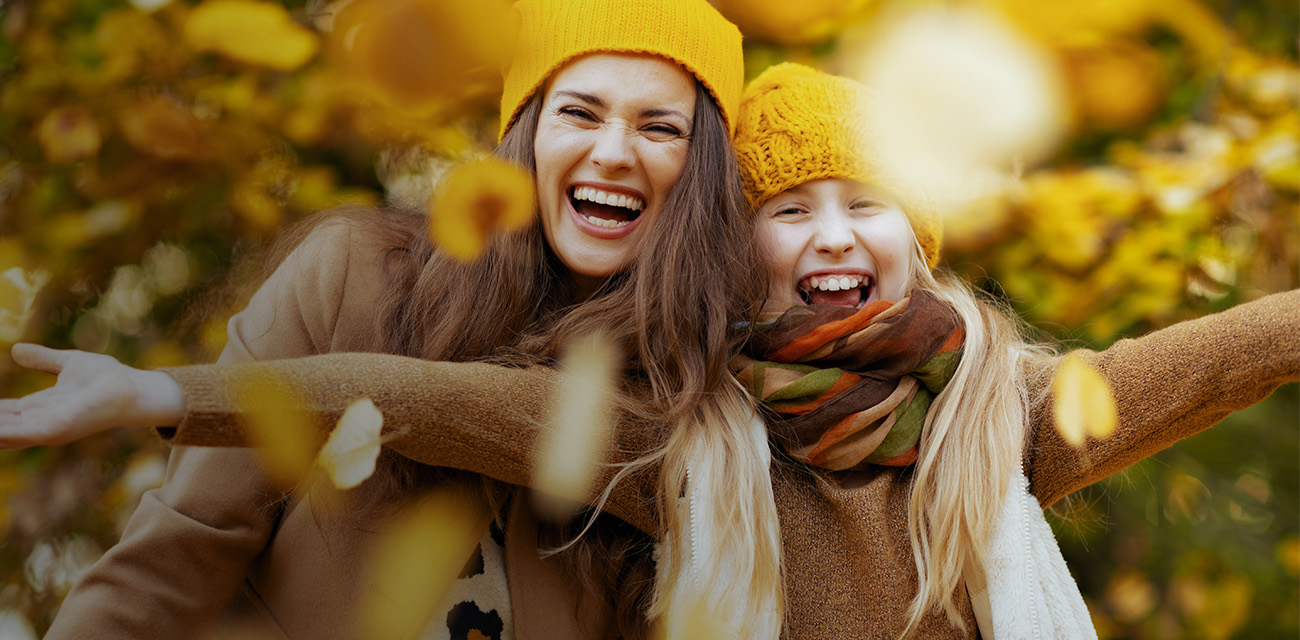 Ein Mädchen und eine Frau stehen in einem herbstlichen Wald. Beide haben gelbe Hauben auf und lachen in die Kamera.